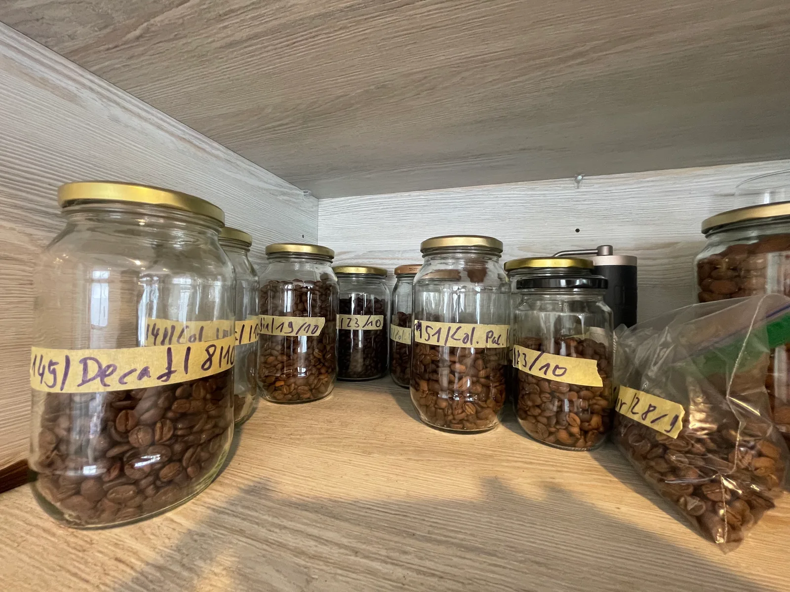 A cupboard shelf of glass jars of home-roasted coffee, each labelled with a batch number and date on masking tape