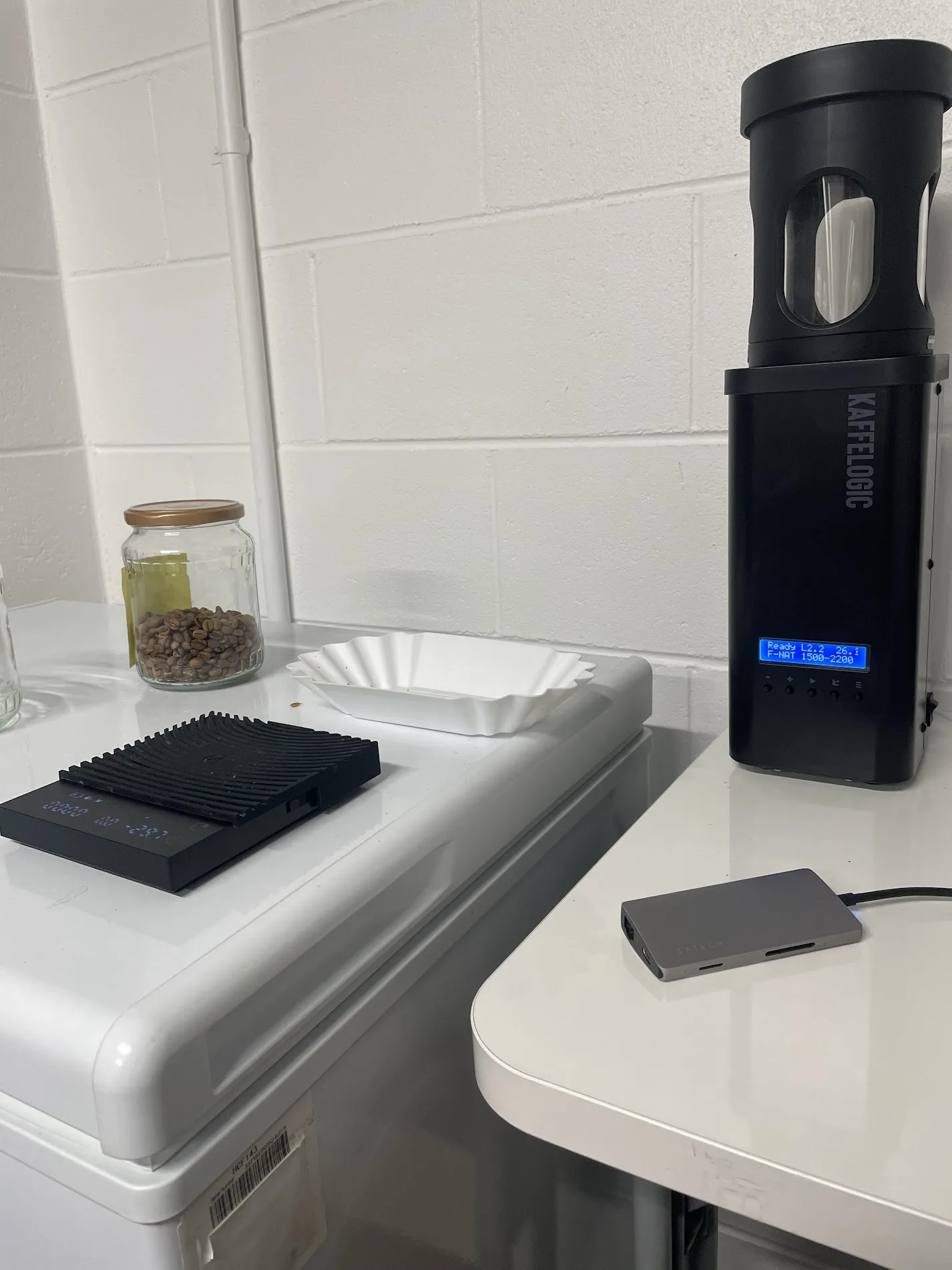 A Kaffelogic Nano roaster on the bench next to a scale, a cooling tray and a jar of green beans